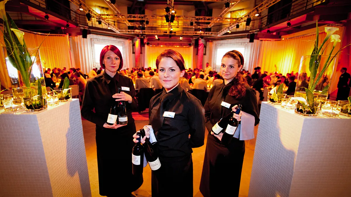 Drei Servicemitarbeiterinnen mit Getränkeflaschen beim Gala Dinner im OKTOGON auf Zollverein in Essen, vor festlich beleuchtetem Bankettsaal