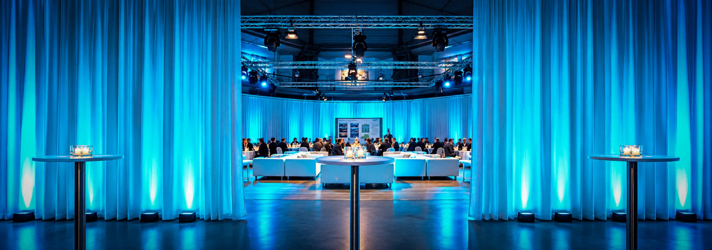 Blick durch blau beleuchtete Vorhänge auf eine Lounge-Veranstaltung mit Gästen und weißen Sofas im OKTOGON auf Zollverein in Essen