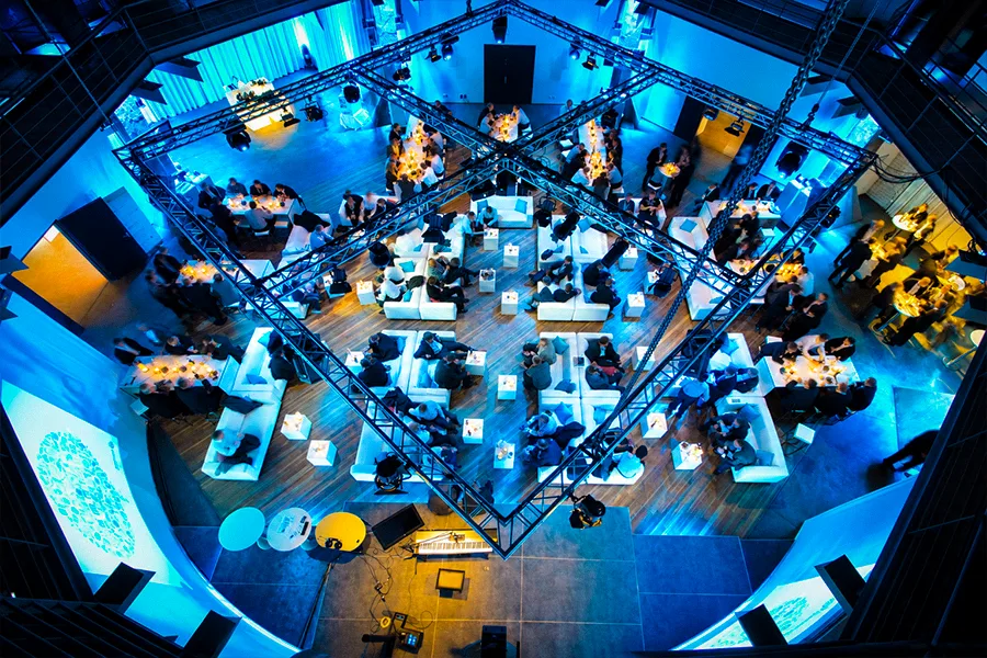 Vogelperspektive auf ein Get-together mit Gästen, weißen Lounge-Sofas und Stehtischen bei blauer Eventbeleuchtung im OKTOGON auf Zollverein in Essen