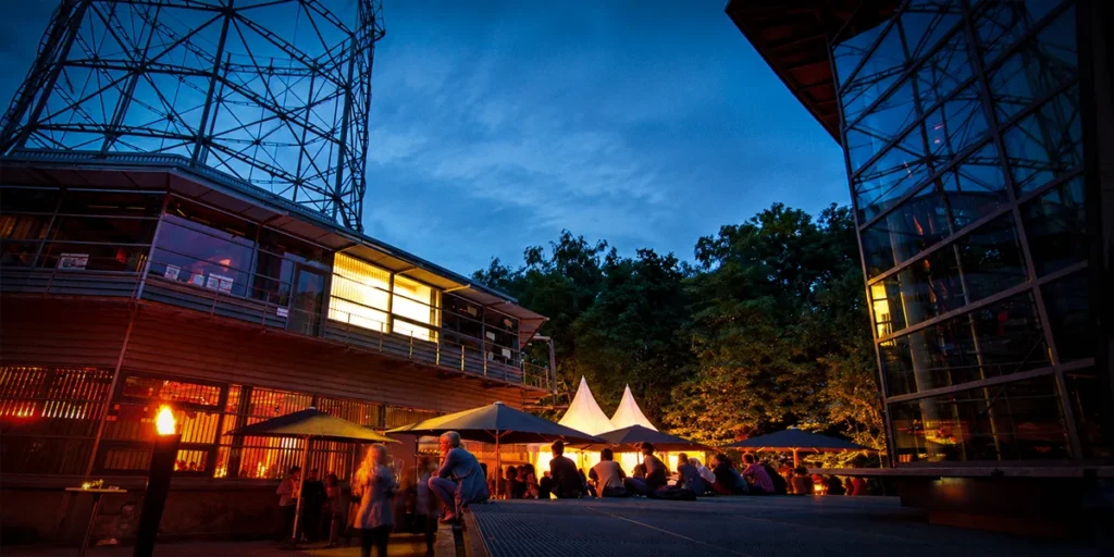 Get-together im Außenbereich des OKTOGON auf Zollverein in Essen mit Gästen, Sonnenschirmen und beleuchteten Zelten in der Abenddämmerung